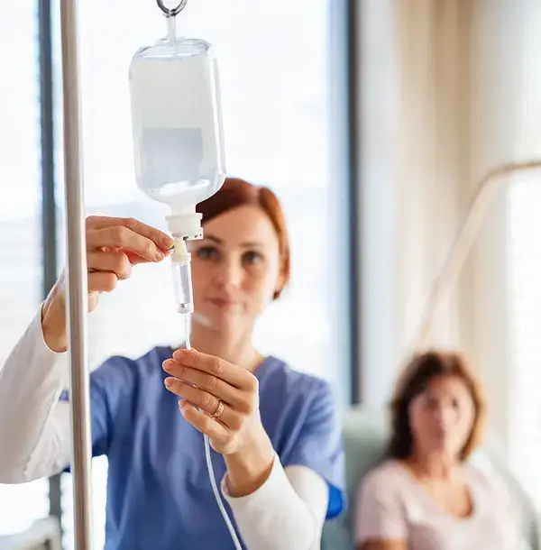 Nurse adjusting an IV drip in a medical setting, patient in background.